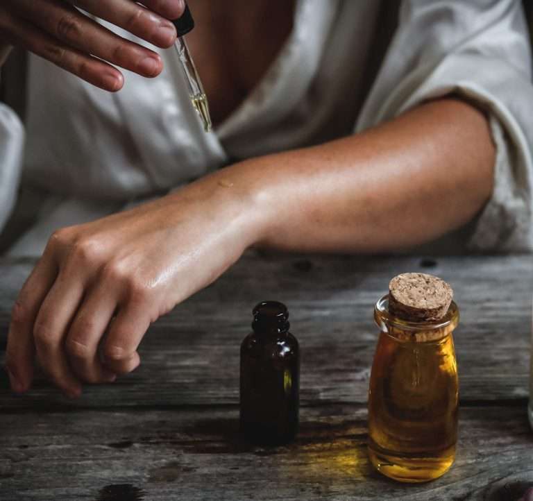 Boccette di oleoliti appoggiati su un tavolo in legno rustico. Sullo sfondo dettaglio del braccio e della mano di una persona che sta versando l'loeolito sulla pelle con un contagocce di vetro
