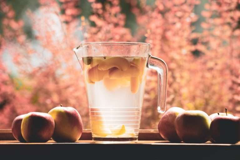 Immagine di una brocca di vetro con infusi di frutta appoggiata su un tavolo con accanto mele e sullo sfondo in dissolvenza fiori a stelo di colo rosa scura che vira all'arancio e al gialo
