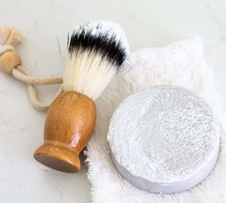 Pennello da barba con manico in legno color miele accanto a una cialda grigio chiaro di crema per depilazione appoggiati su asciugamano bianco. Il tutto su sfondo neutro: gli strumenti neessari per la rasatura