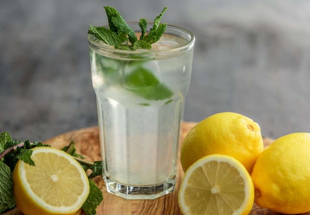 Acqua tiepida e limone con un ciuffo di menta in un alto bicchiere di vetro appoggiato su un tavolo di legno, un ottimo detox di Primavera