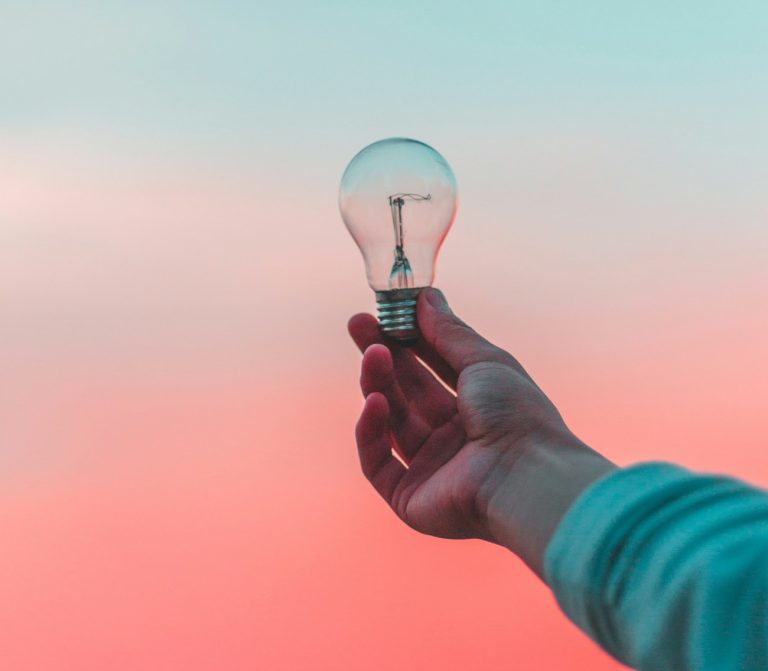 Mano che regge una lampadina su uno sfondo rosa che sfuma nell'azzurro a indicare le risposte alle domande sul benessere naturale
