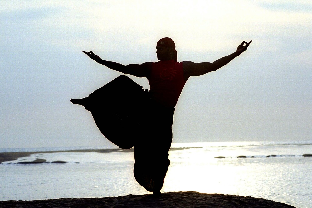 Figura in controluce che pratica ginnastiche orientali in riva al mare al tramonto per prevenire e combattere i dolori articolari